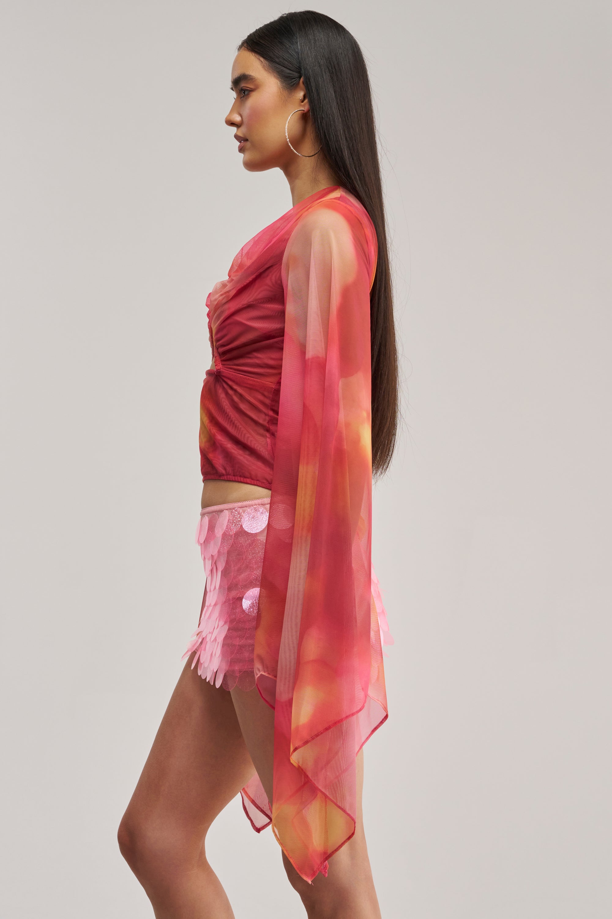 Wearing the CELESTE CUTOUT DRAPE TOP in coral, a woman is shown in profile with flowing sleeves, styled with a pink sequin mini skirt and large hoop earrings. Her long straight hair stands out against a plain light background.