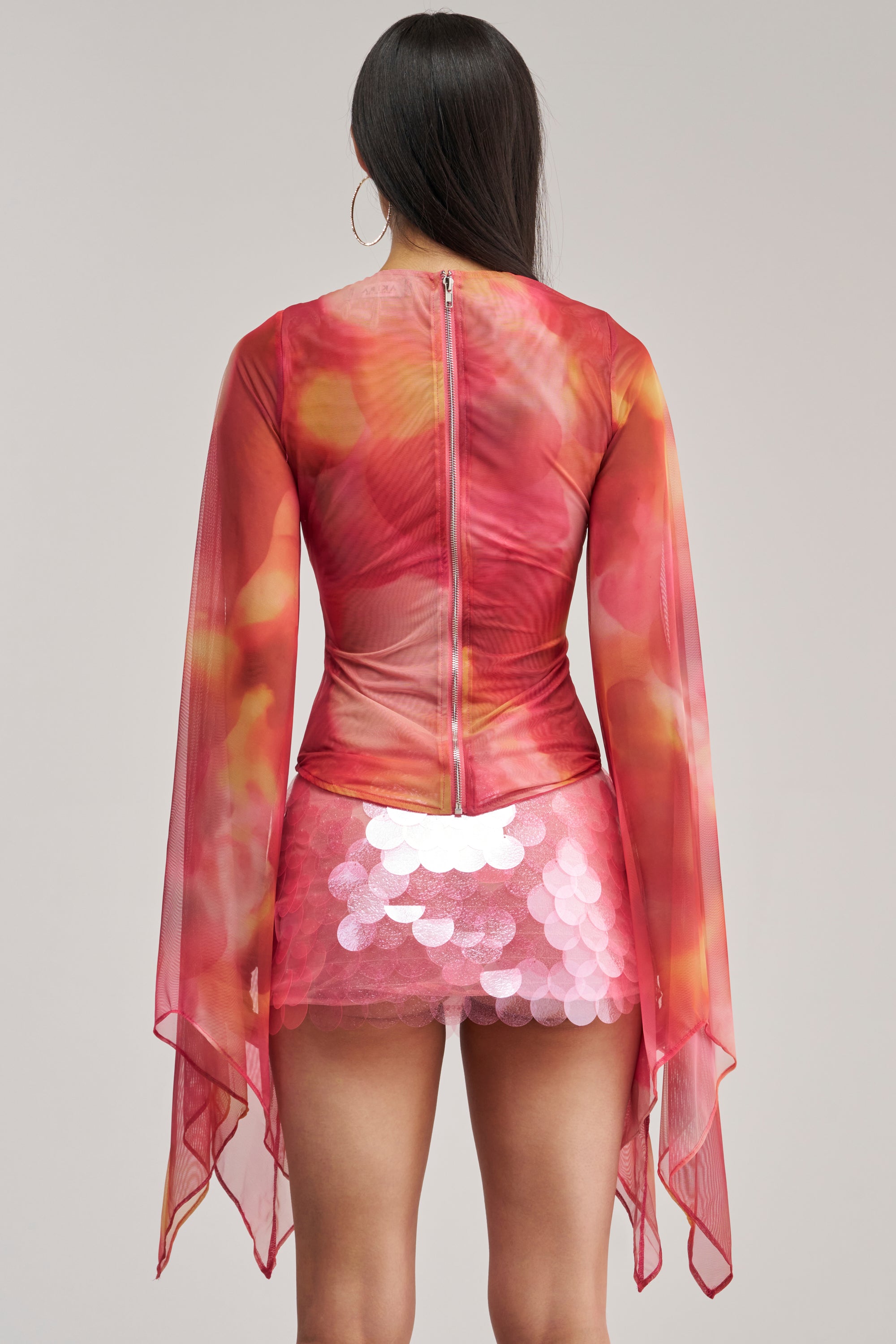 A person with long dark hair is shown from the back, wearing the CELESTE CUTOUT DRAPE TOP—a sheer mesh top in vibrant red and orange hues with flowing sleeves—paired with a short skirt decorated with large, shiny pink sequins.