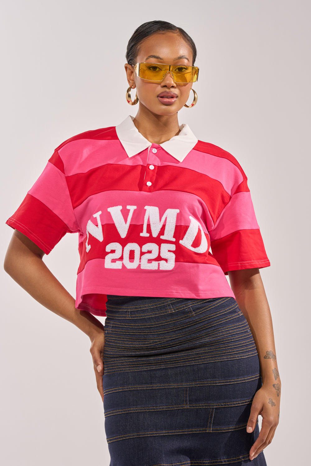 A woman in yellow-tinted sunglasses, a NEVERMIND KNIT TEE with red and pink stripes reading "NVMD 2025," and a dark denim skirt stands confidently against a plain backdrop. 0