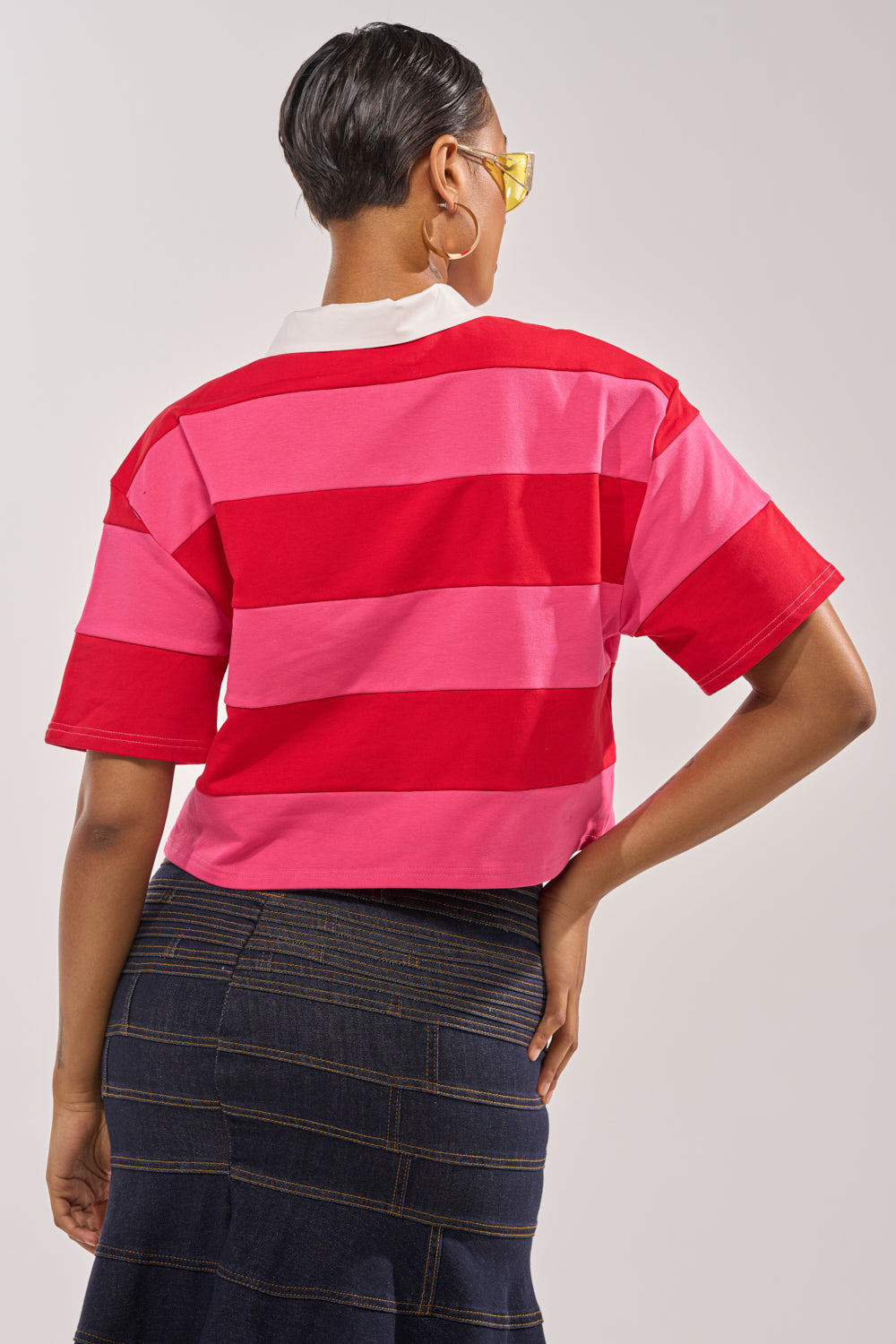 A person with short hair, yellow glasses, and a dark denim skirt stands facing away with one hand on their hip, wearing the NEVERMIND KNIT TEE—a striped red and pink short sleeve top. 3
