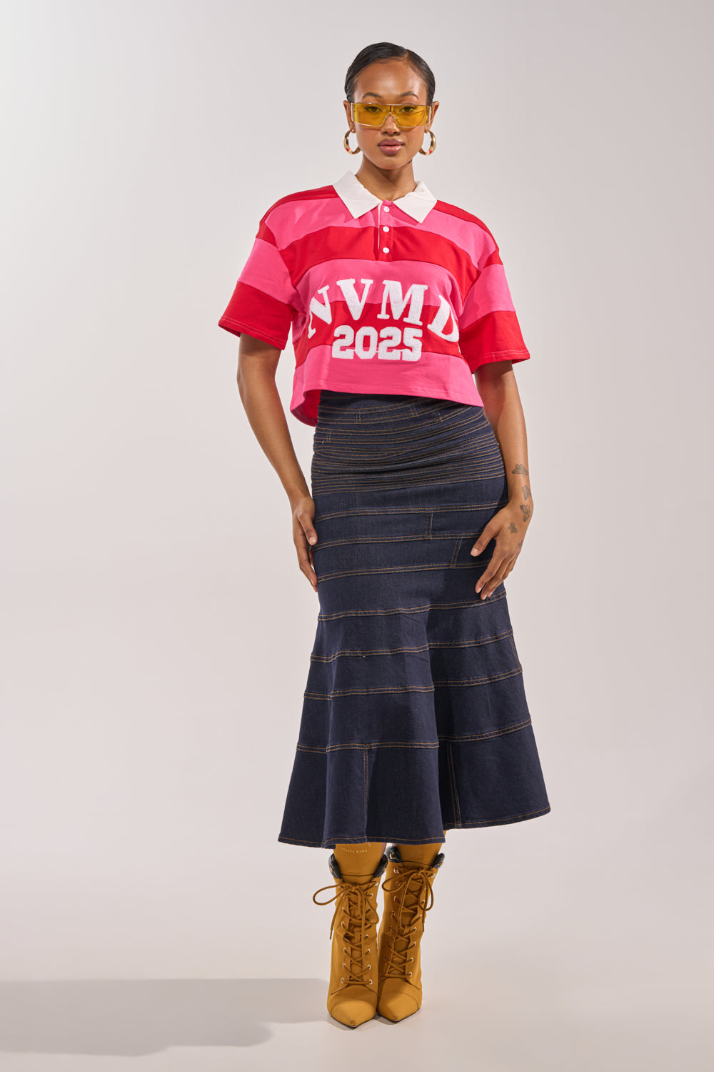 A woman poses against a plain background wearing yellow sunglasses, the NEVERMIND KNIT TEE with "NVMND 2025" print, a long dark denim skirt, and mustard yellow lace-up boots. 4