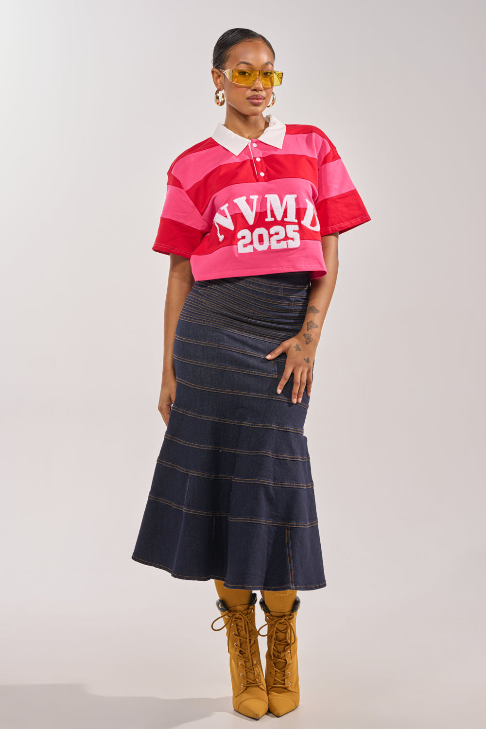 A person poses in the NEVERMIND KNIT TEE with pink and red stripes reading "NVMD 2025," styled with a dark denim skirt, yellow heeled boots, and yellow-tinted sunglasses against a plain background. 5