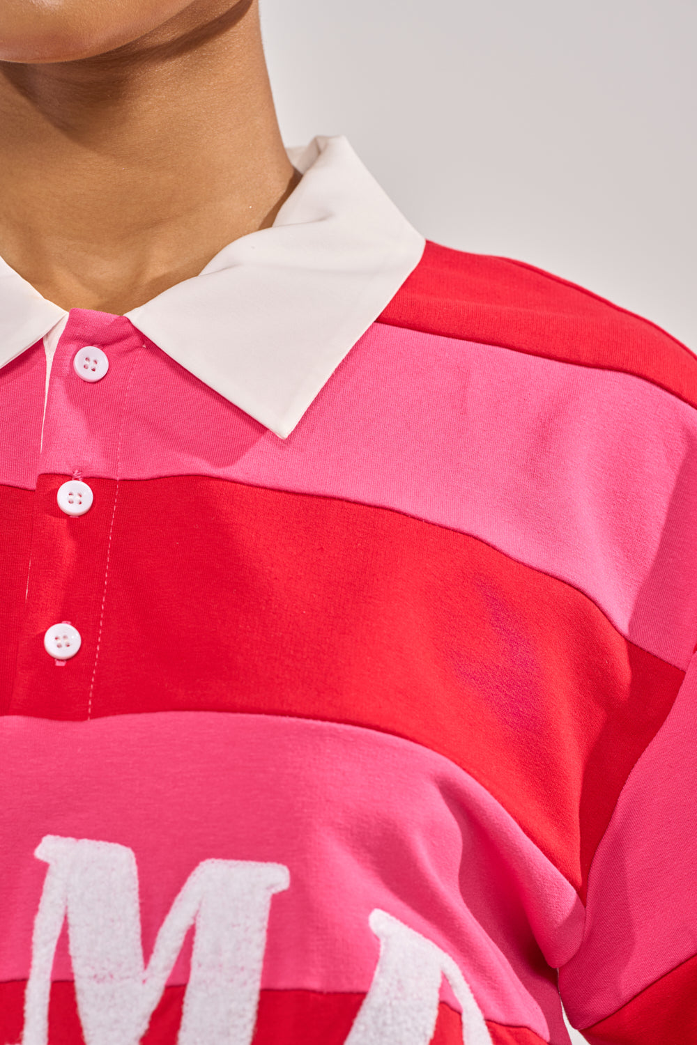 A close-up of a person wearing the NEVERMIND KNIT TEE, a red and pink striped short sleeve top with white buttons and a bold white “M” on the chest for a classic varsity style. 6
