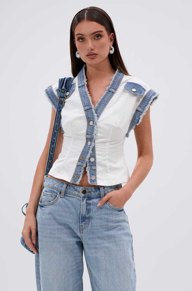 A woman with long brown hair models the DENIM BABY BUTTON DOWN TOP, paired with light blue jeans, hoop earrings, and a crossbody bag. She poses in street-style fashion, hand in pocket against a plain background. 0