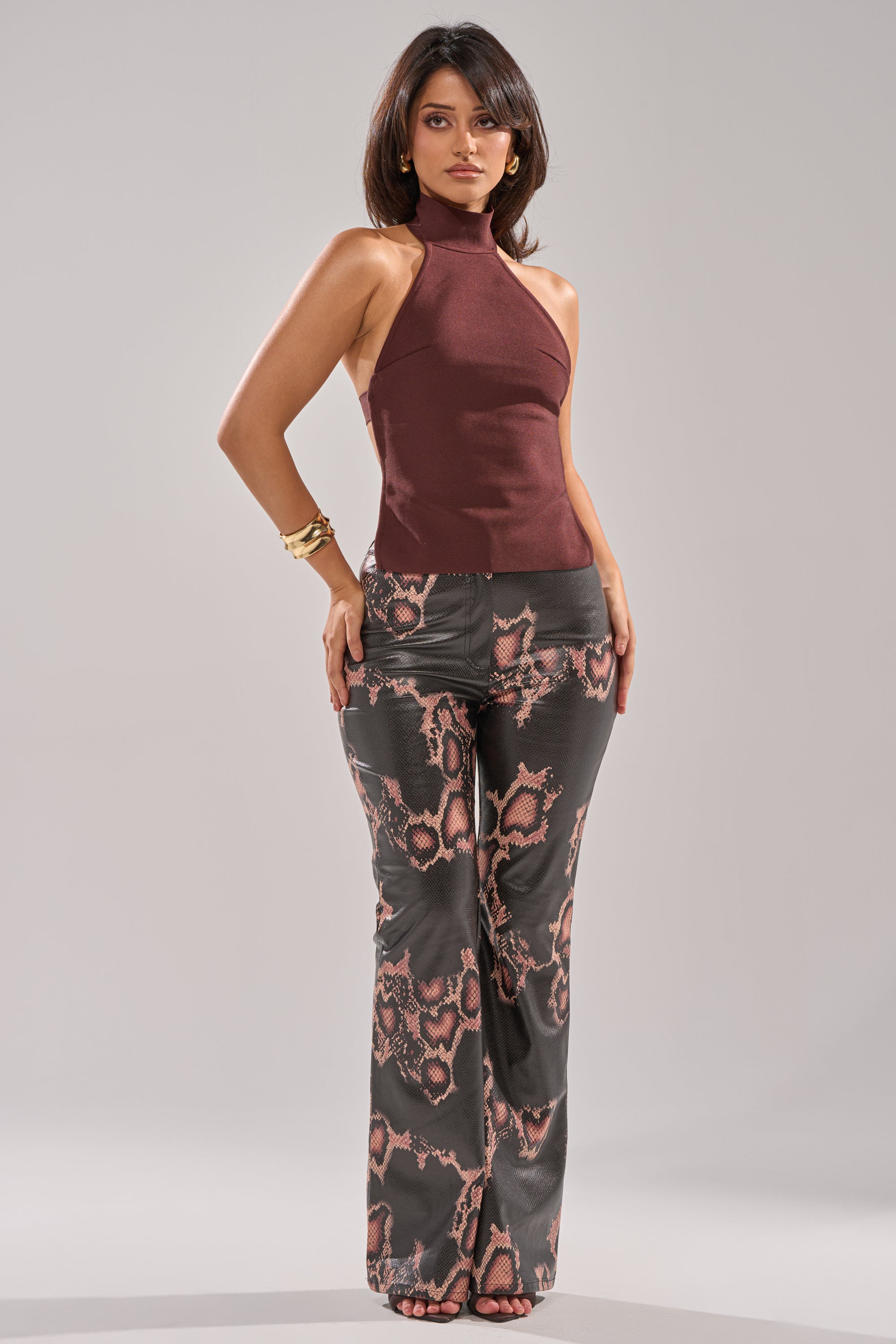 A woman with shoulder-length dark hair and gold bracelets poses in the TRY ME BANDAGE BLOUSE, a sleeveless maroon halter top featuring an open-back detail, paired with high-waisted black pants with a pink and brown snakeskin pattern. 4