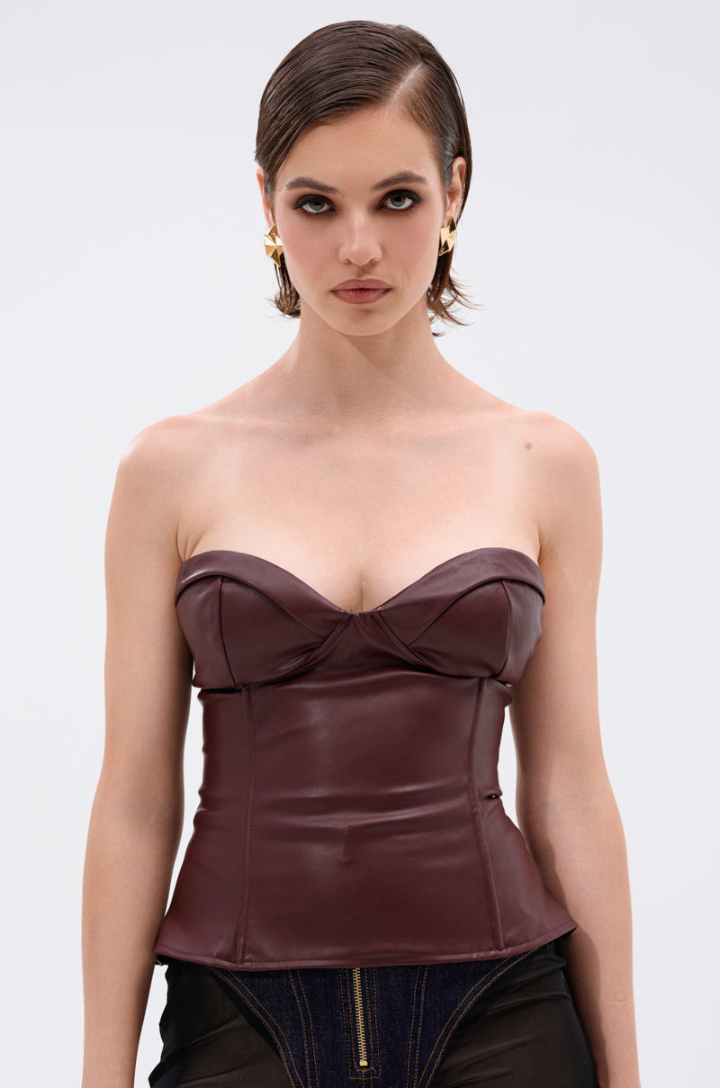 A woman with short, slicked-back hair wears the WHATEVER SHE WANT FAUX LEATHER CORSET in burgundy and gold earrings. She stands against a plain light background, looking directly at the camera with a neutral expression.