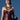 A woman with short dark hair and a serious look stands against a blue background, holding a black balloon animal while wearing the WHATEVER SHE WANT FAUX LEATHER CORSET and layered gold necklaces.