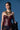 A woman with short dark hair and a serious look stands against a blue background, holding a black balloon animal while wearing the WHATEVER SHE WANT FAUX LEATHER CORSET and layered gold necklaces.