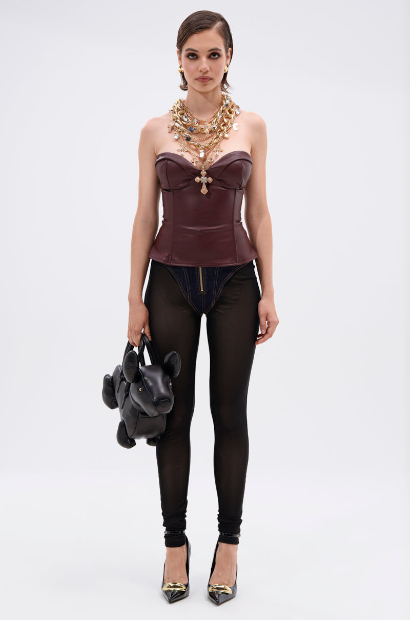 A woman stands against a plain background wearing the WHATEVER SHE WANT FAUX LEATHER CORSET paired with sheer black leggings over denim shorts, black heels, layered gold necklaces, and holding a playful dog-shaped black handbag.