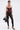 A woman poses against a plain background wearing the WHATEVER SHE WANT FAUX LEATHER CORSET, sheer black leggings, pointed heels, layered gold jewelry, and holding a dog-shaped black handbag.