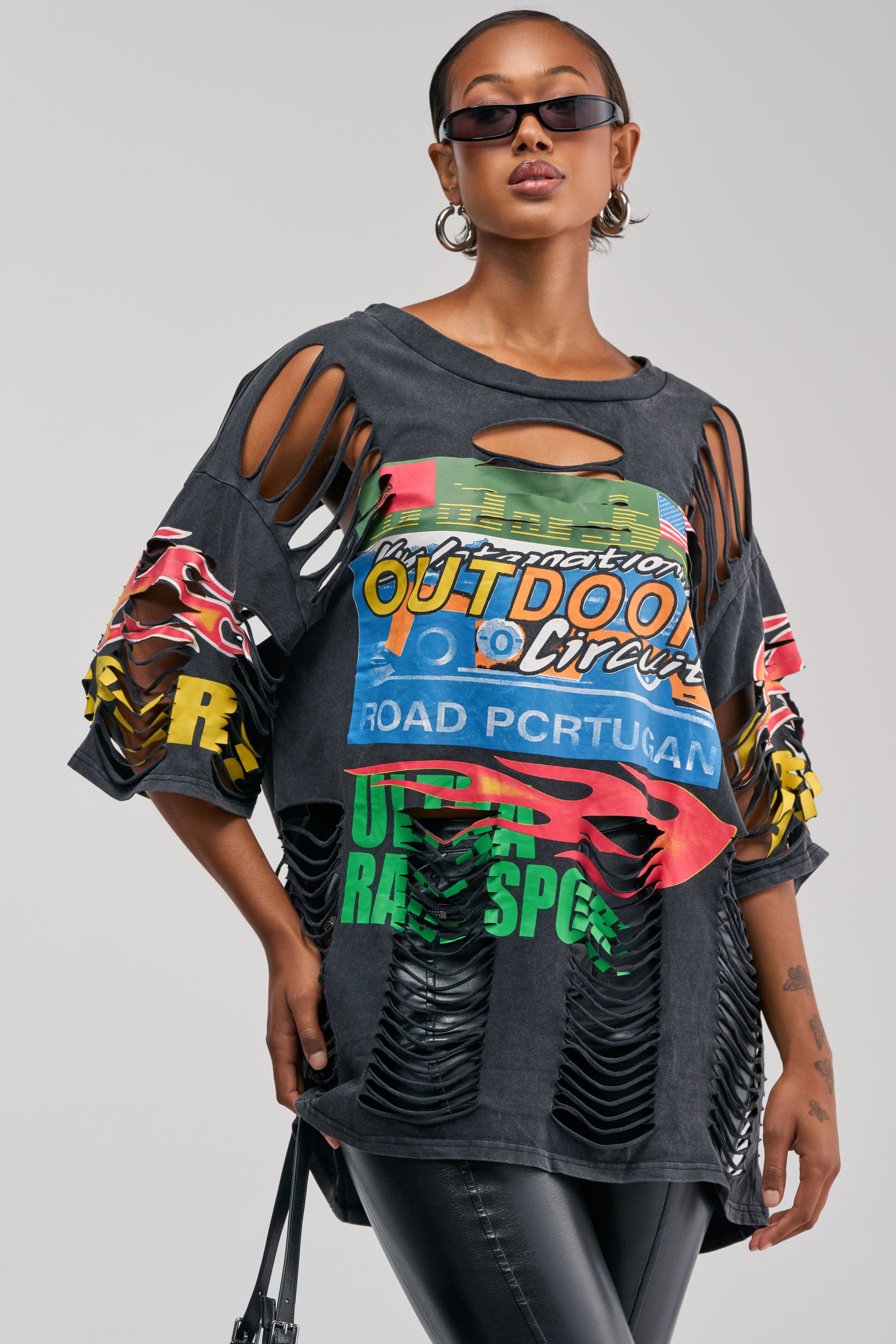 A woman wearing sunglasses and hoop earrings styles the OFF TO THE RACES DISTRESSED OVERSIZED TEE, featuring colorful prints and shredded details on the sleeves and torso, with black pants for a bold streetwear look.
