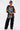 Wearing large sunglasses, furry black pants, and carrying an AKIRA Label bag with toy cars, a person stands confidently in the OFF TO THE RACES DISTRESSED OVERSIZED TEE with racing graphics against a plain white background.
