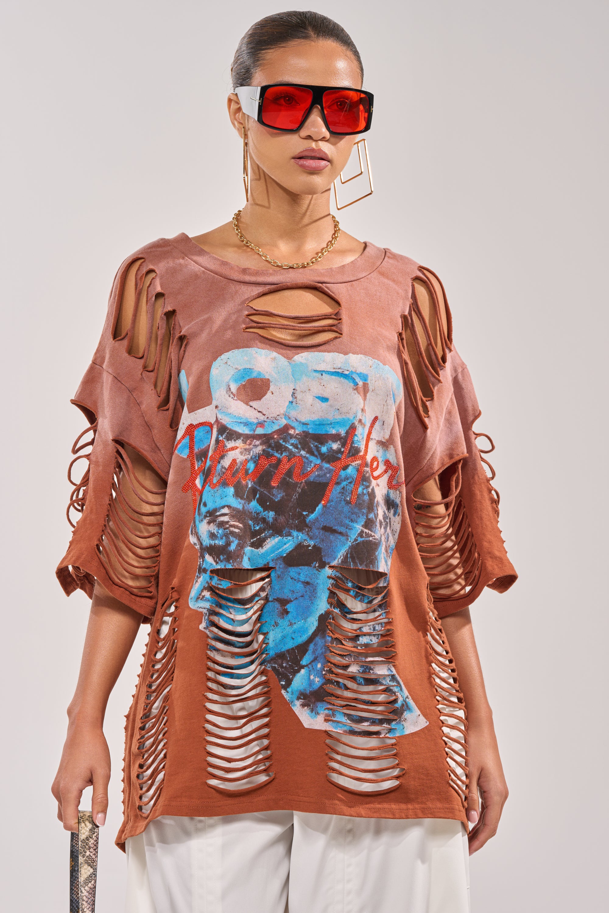 A woman in oversized sunglasses and geometric earrings models the BYE GIRL OVERSIZED DISTRESSED TEE IN BROWN—a streetwear t-shirt with horizontal cutouts, blue graphic print, paired with white pants and a gold chain necklace. 0
