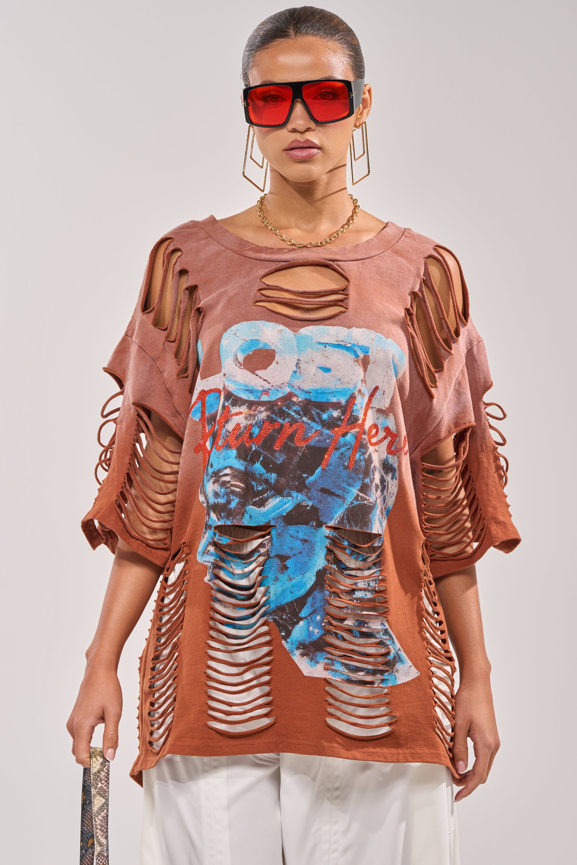 A woman wearing red sunglasses, gold jewelry, white pants, and the BYE GIRL OVERSIZED DISTRESSED TEE IN BROWN with blue and red graphics and shredded cut-outs stands against a plain background. 1