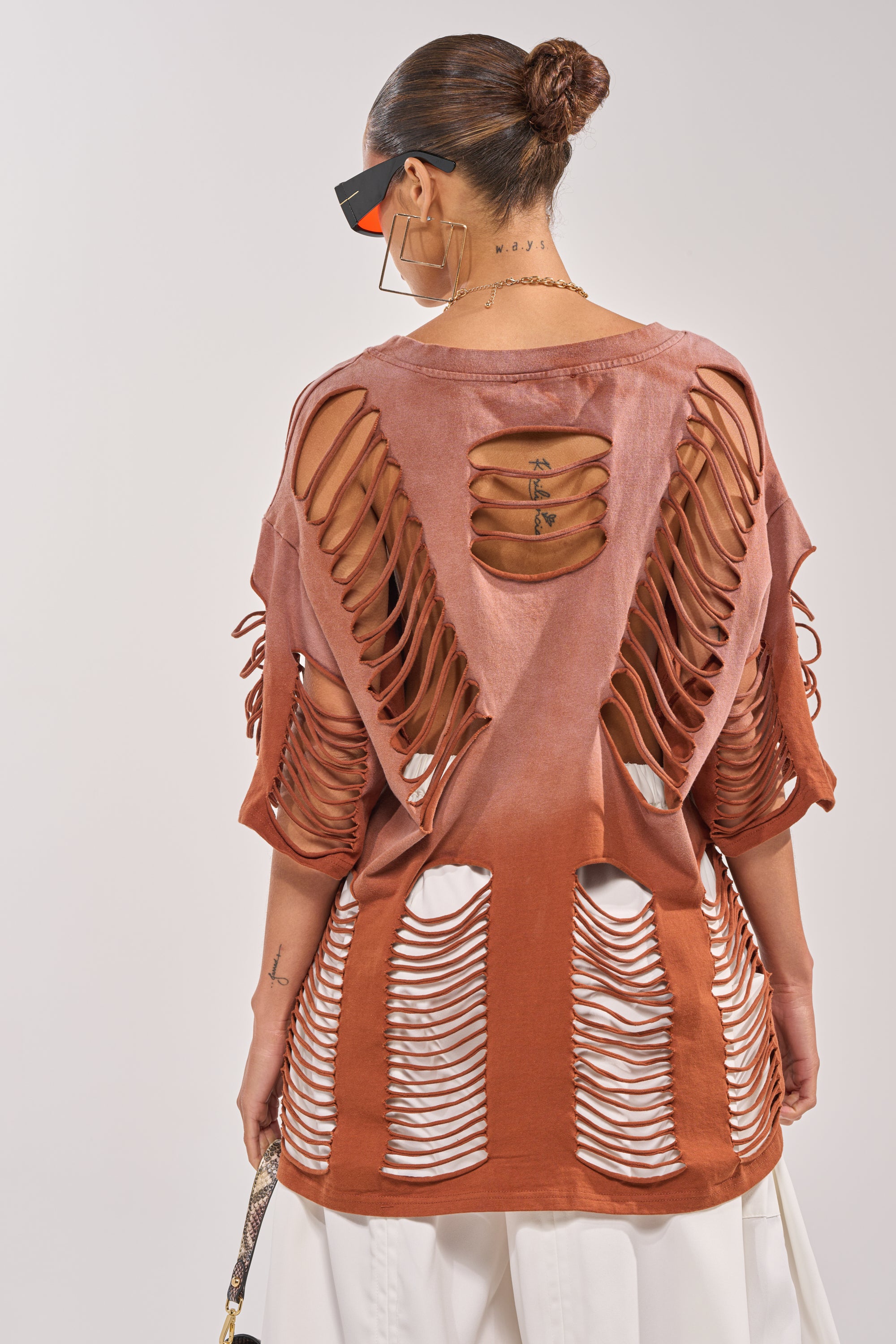 A woman with her hair in a bun, wearing geometric earrings and sunglasses, is seen from behind in the BYE GIRL OVERSIZED DISTRESSED TEE IN BROWN over a white top and loose white pants. 4
