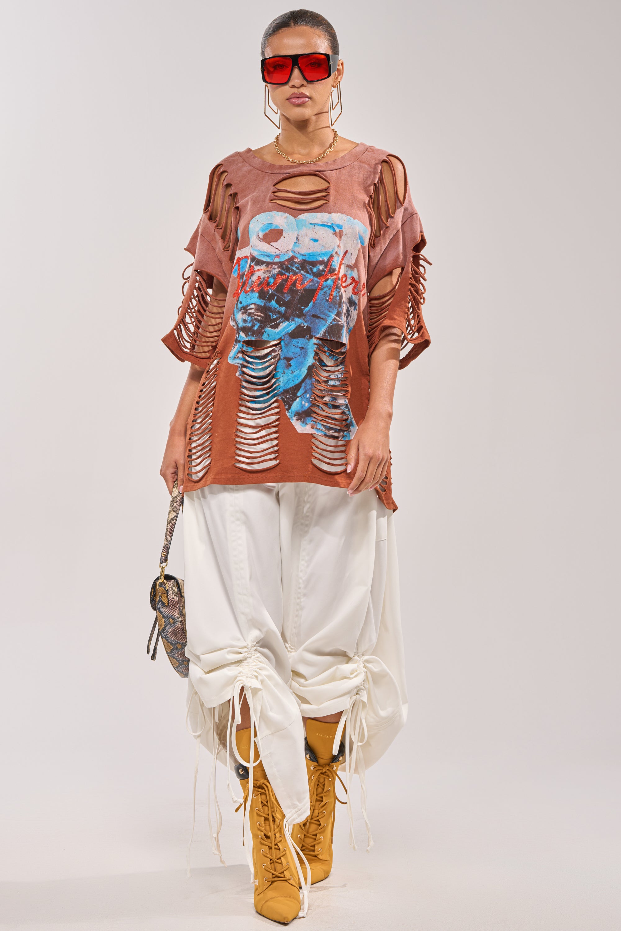 A woman wears oversized red sunglasses, the BYE GIRL OVERSIZED DISTRESSED TEE IN BROWN, loose white pants tied at the ankles, tan lace-up boots, gold hoop earrings, and carries a small patterned bag. 5