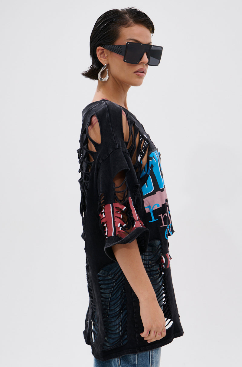 Wearing large square sunglasses, hoop earrings, and the BYE GIRL OVERSIZED DISTRESSED TEE with colorful graphics and multiple rips, a person stands in profile against a white background, showcasing bold edge. 2