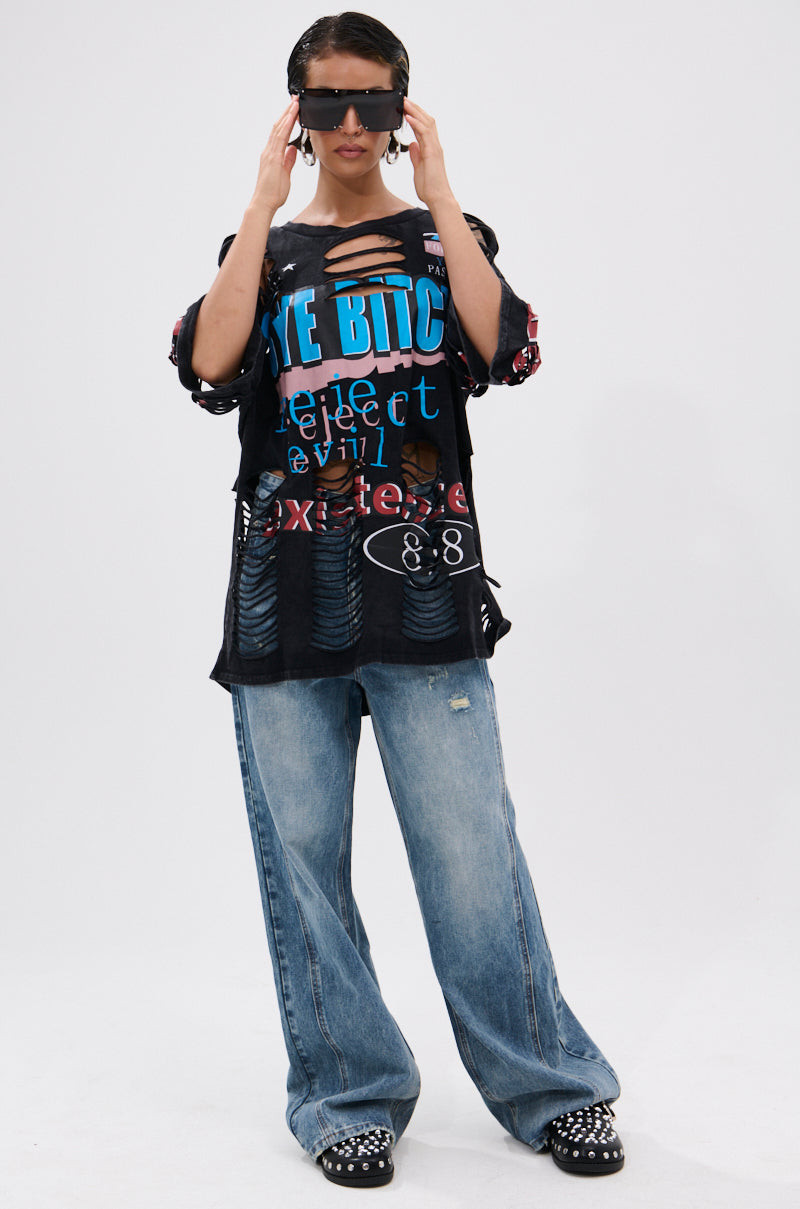 Wearing large sunglasses, the model showcases the BYE GIRL OVERSIZED DISTRESSED TEE with a vintage graphic, paired with loose blue jeans and studded black shoes, standing against a white background and holding the sunglasses for a bold look. 4