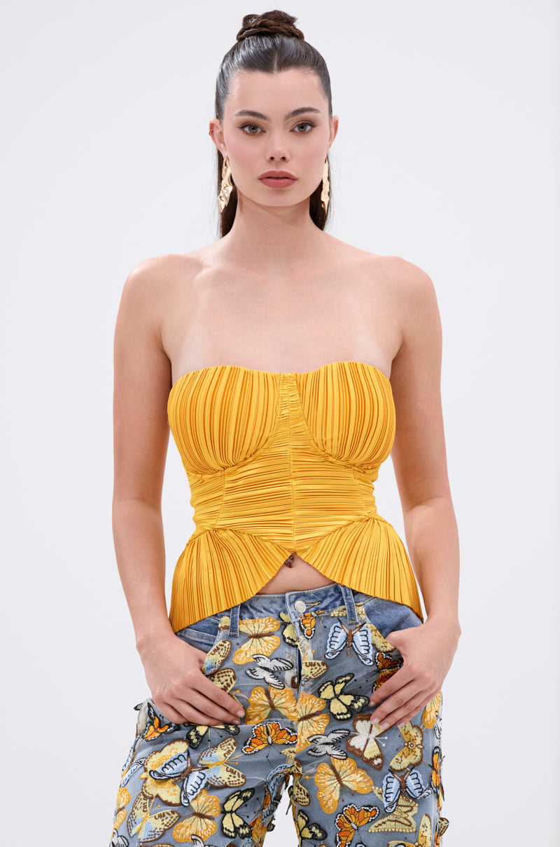 A woman with long brown hair in a high ponytail wears the KEEP GOING PLISSE CORSET IN YELLOW and blue jeans with colorful butterfly prints, standing confidently with her hands in her pockets against a plain background.