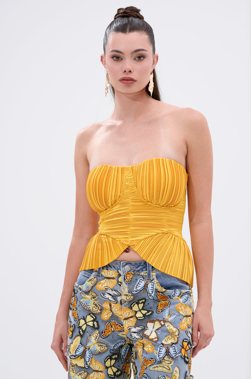 A woman with long brown hair in a high ponytail wears the KEEP GOING PLISSE CORSET IN YELLOW and blue jeans with colorful butterfly prints, standing against a plain white background.