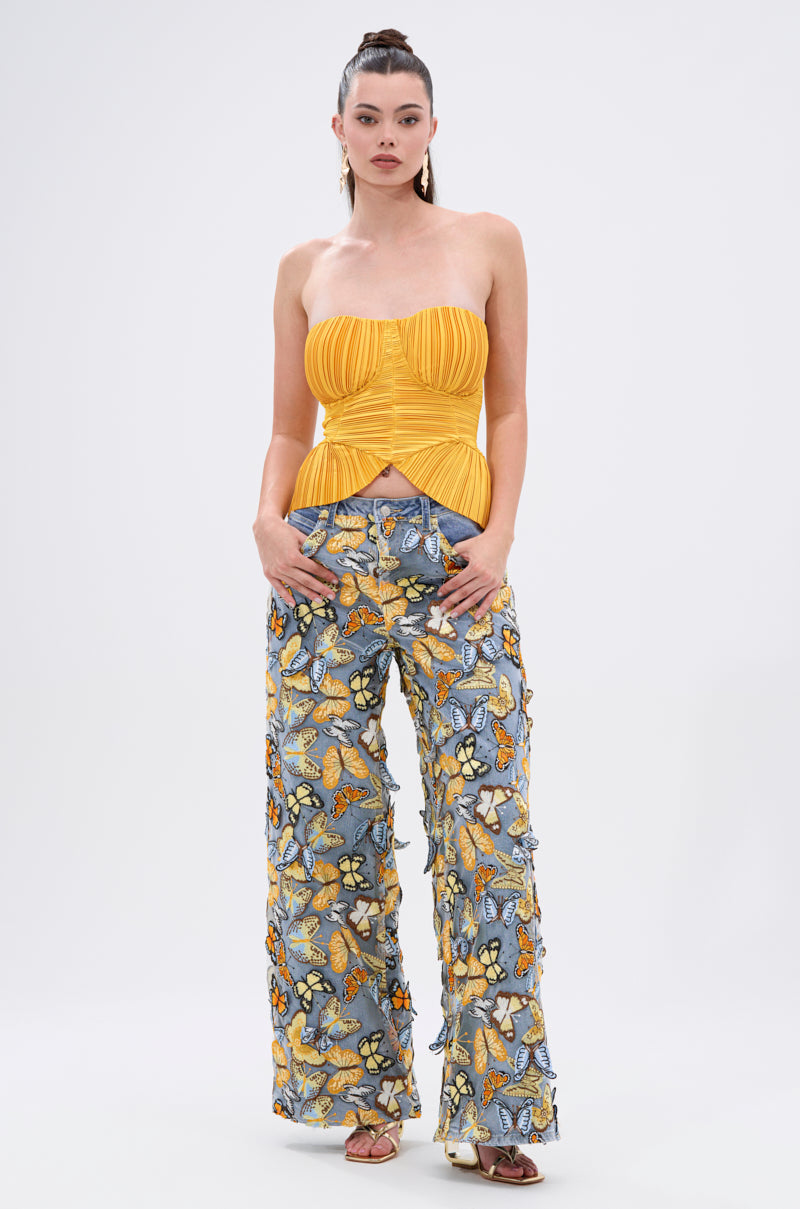 Woman stands against a plain background, wearing the KEEP GOING PLISSE CORSET IN YELLOW and high-waisted blue jeans with colorful butterfly patterns, finished with strappy heels.