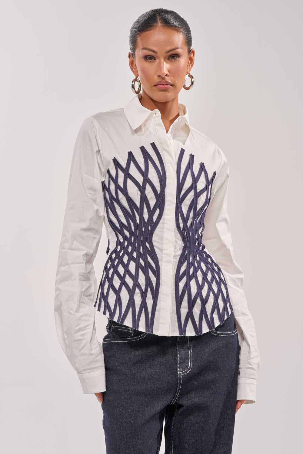 A woman wears the ON A NEW LEVEL POPLIN BUTTONDOWN, a white blouse with a sculptural black geometric design, paired with dark jeans. Slicked-back hair and large hoop earrings add to her bold, modern style. 0