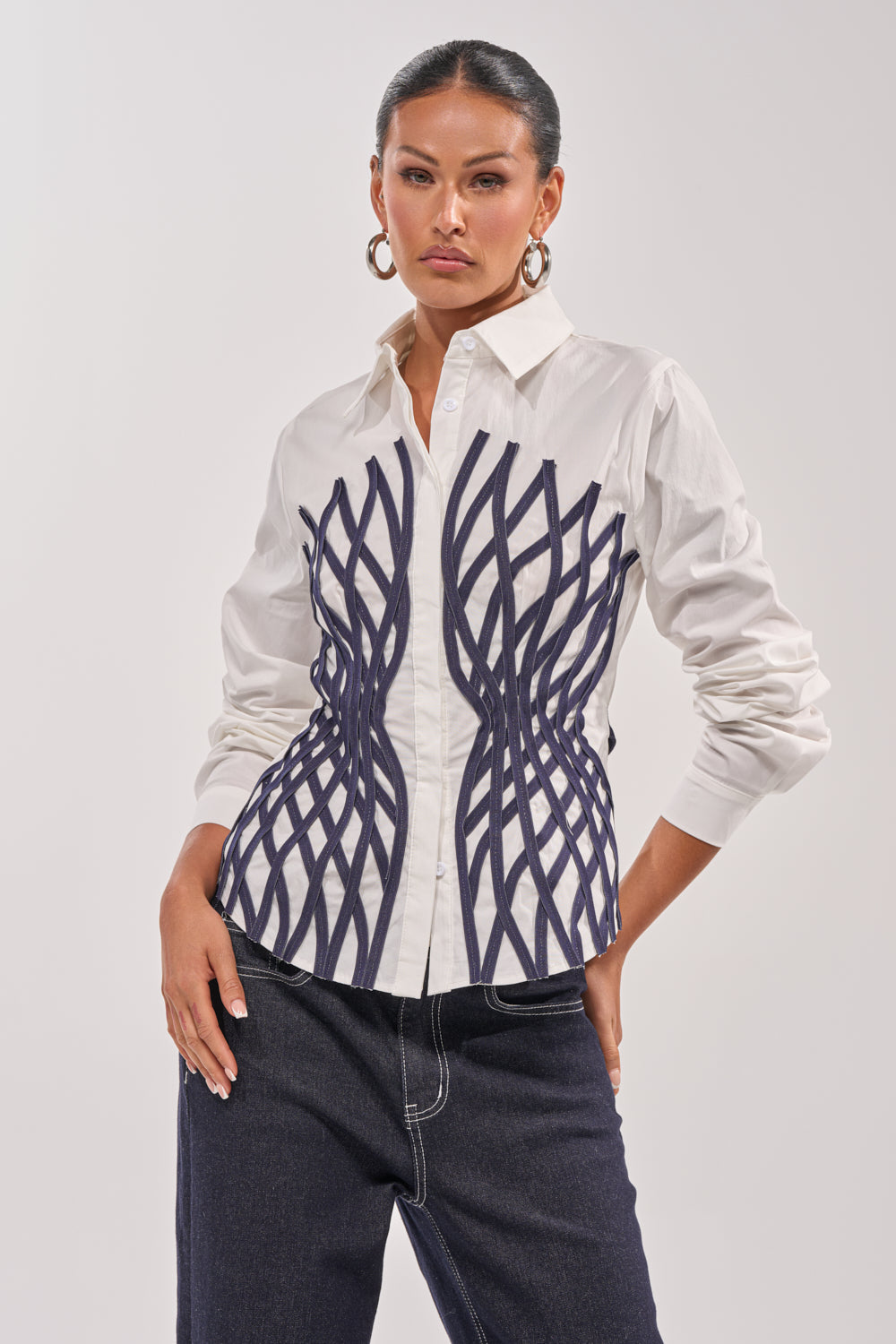 A woman models the ON A NEW LEVEL POPLIN BUTTONDOWN, a white blouse with black geometric lines in a corset-inspired pattern, paired with dark jeans. She accessorizes with hoop earrings and has her hair pulled back against a plain light background. 1