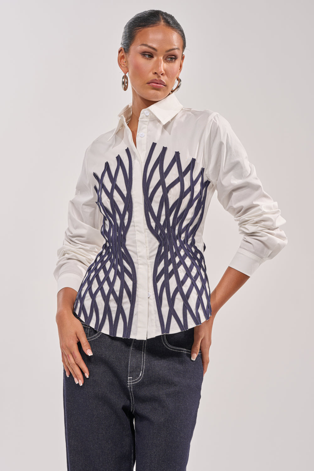 A woman stands against a light background, wearing the ON A NEW LEVEL POPLIN BUTTONDOWN—a structured, long sleeve white blouse with a dark wavy geometric pattern on the front—paired with dark jeans. She has hoop earrings and her hair is pulled back. 2