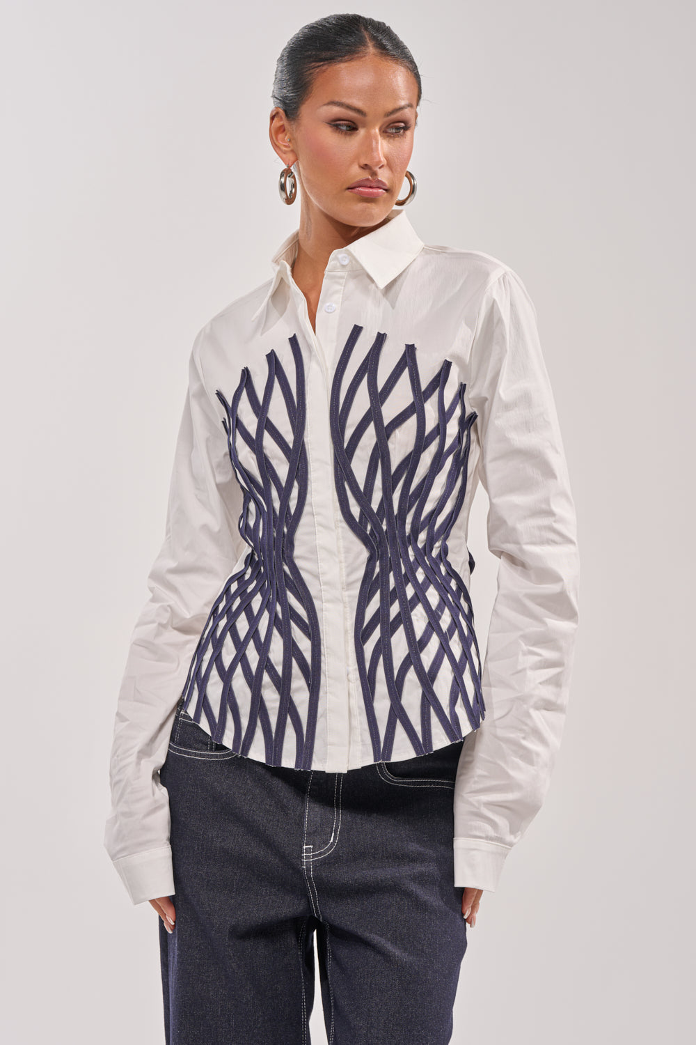 A woman wears the ON A NEW LEVEL POPLIN BUTTONDOWN, a fitted white blouse with black wavy vertical lines, paired with dark jeans. She accessorizes with hoop earrings, her hair pulled back, and stands against a plain light background. 3