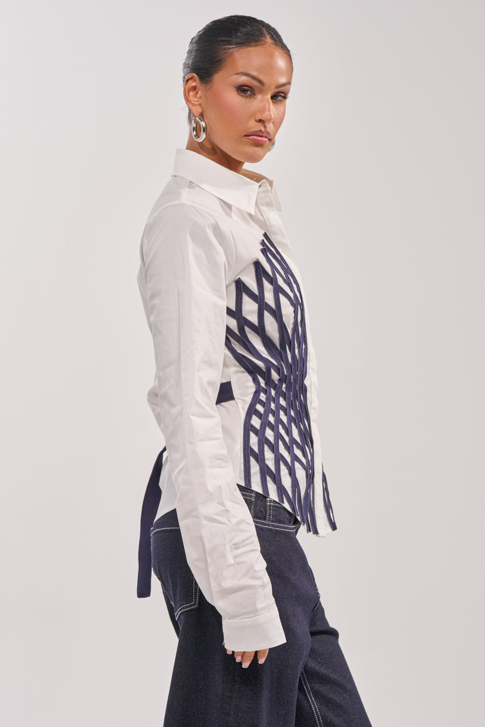 A woman with dark hair in a ponytail models the ON A NEW LEVEL POPLIN BUTTONDOWN featuring white fabric with navy blue geometric detailing, paired with dark blue pants and hoop earrings, against a plain light background. 4