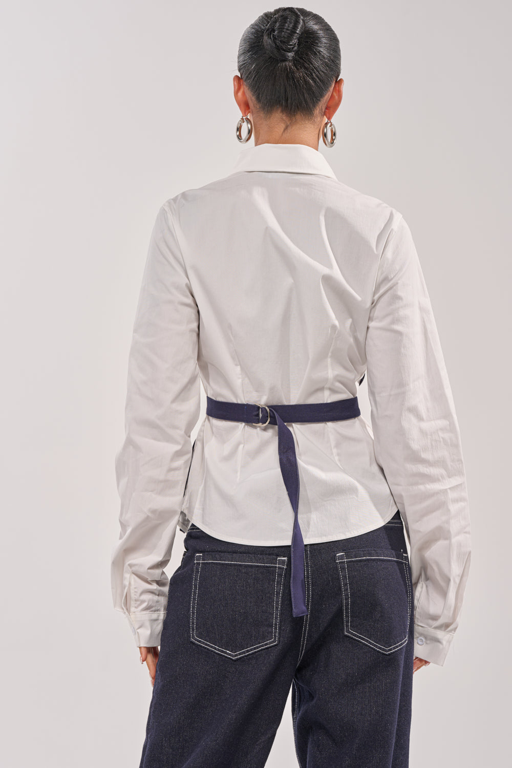Someone stands facing away, wearing the ON A NEW LEVEL POPLIN BUTTONDOWN—a structured white blouse with long sleeves—paired with dark blue jeans featuring visible stitching, a navy tie-back belt, silver hoop earrings, and a neat bun. 5