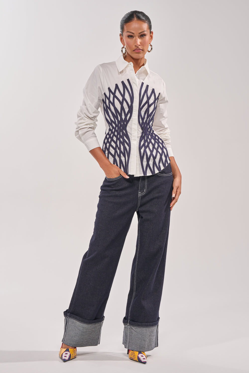 A woman stands confidently in the ON A NEW LEVEL POPLIN BUTTONDOWN, a white blouse with navy geometric waist patterns, paired with wide-leg dark cuffed jeans, colorful pointed heels, and statement earrings against a light background. 6