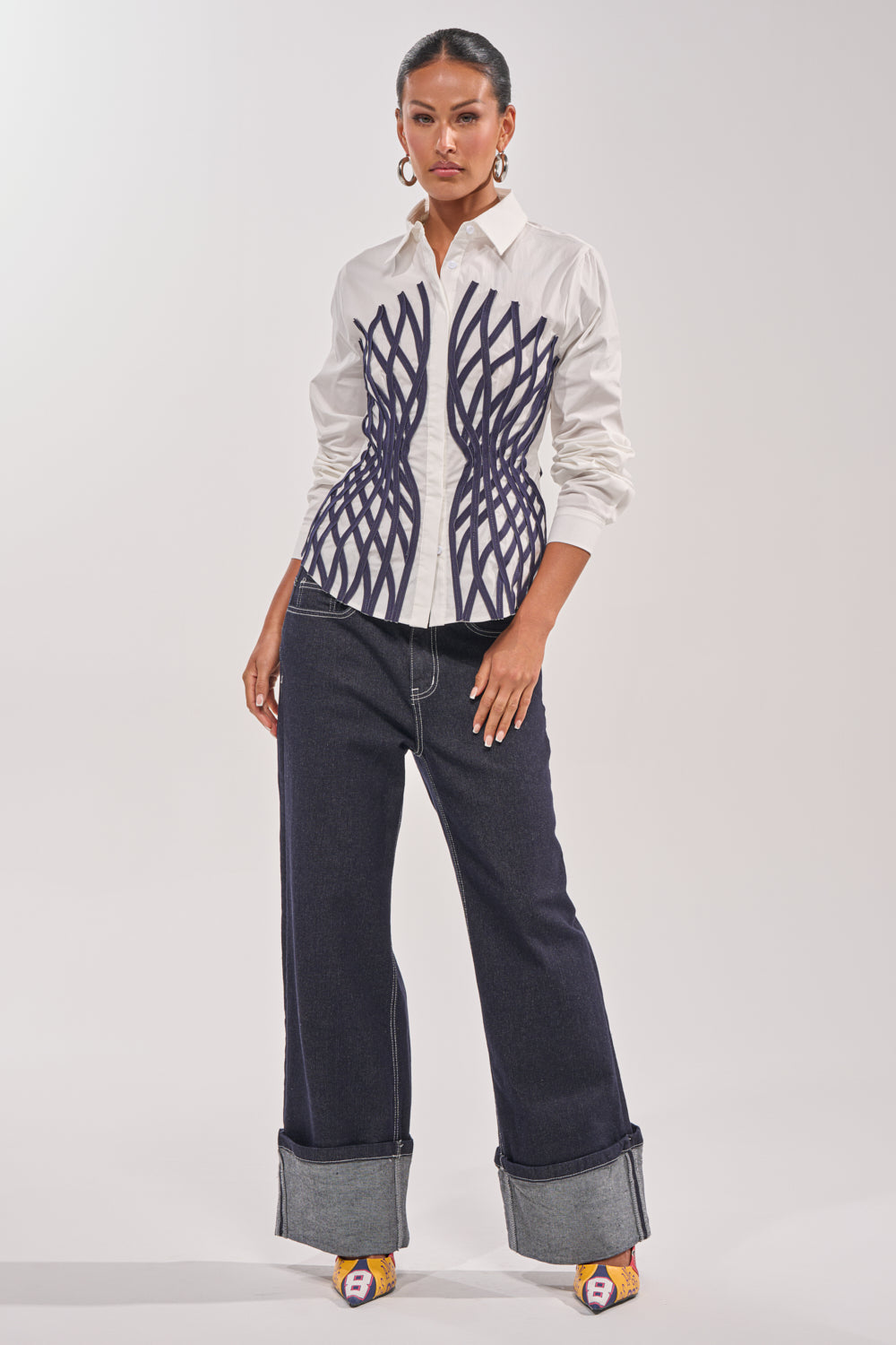 A woman stands confidently in the ON A NEW LEVEL POPLIN BUTTONDOWN, a white blouse with navy geometric waist details, paired with wide-leg dark cuffed jeans, yellow open-toe heels, large hoop earrings, and her hair pulled back. 7