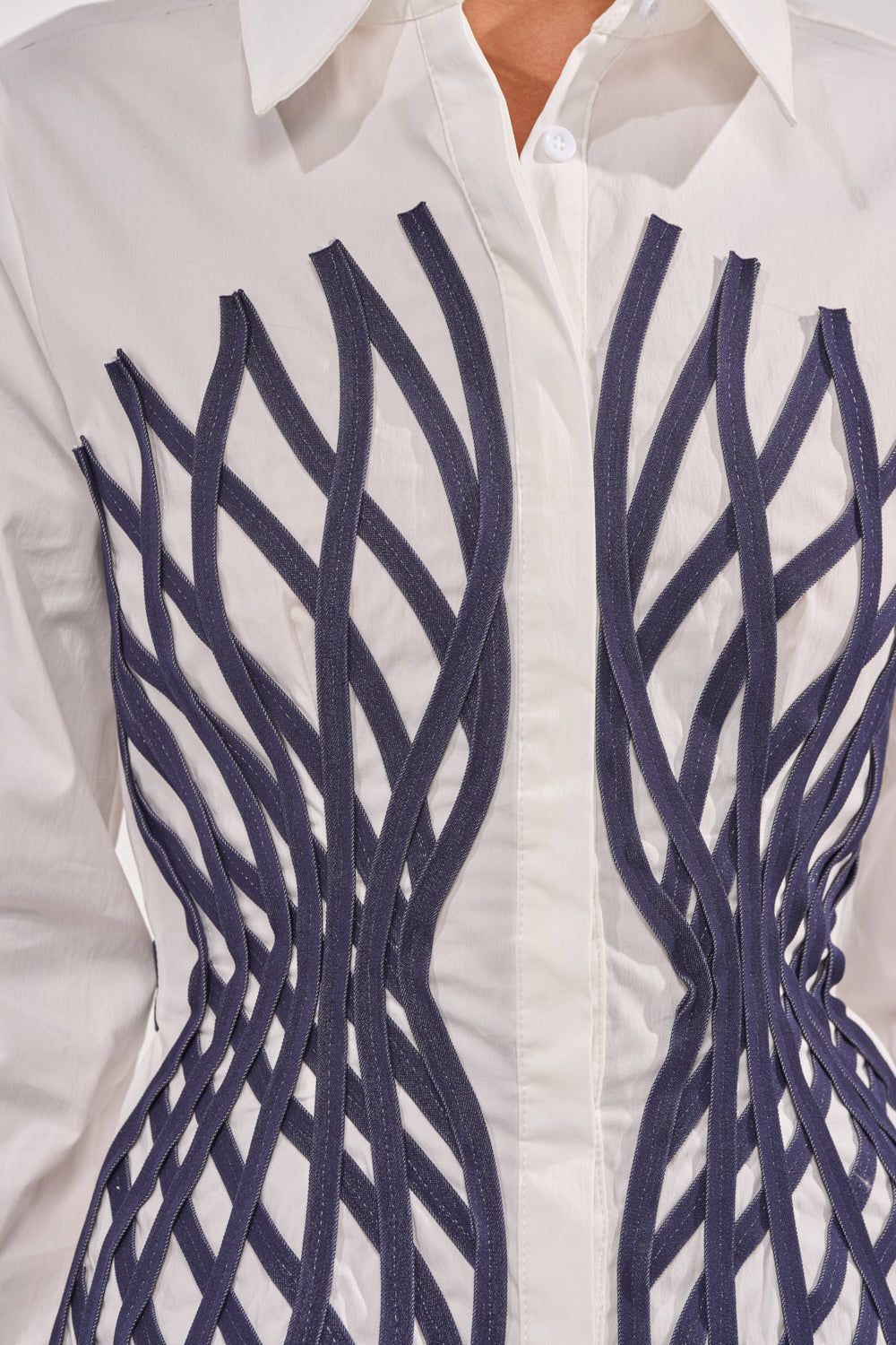 A close-up of the ON A NEW LEVEL POPLIN BUTTONDOWN shows a structured white long sleeve blouse with dark blue, wavy, ribbon-like strips sewn in geometric patterns across the chest and waist for a sculptural effect. 8