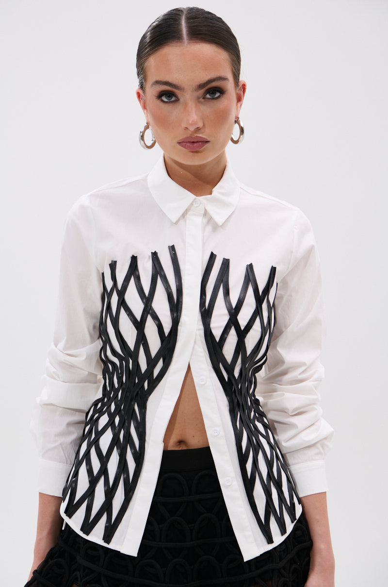 A woman in the ALL OVER THE PLACE BUTTON DOWN—a white tailored shirt with black geometric lattice details and rolled-up sleeves—paired with a black skirt, faux leather waistbelt, hoop earrings, and sleek hair stands against a white background.