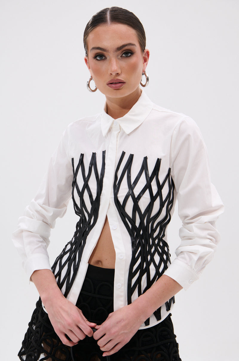 A woman models the ALL OVER THE PLACE BUTTON DOWN, a white shirt with black lattice-like overlays and a tailored fit, paired with black patterned shorts. She sports slicked-back hair, hoop earrings, and bold makeup against a white background.