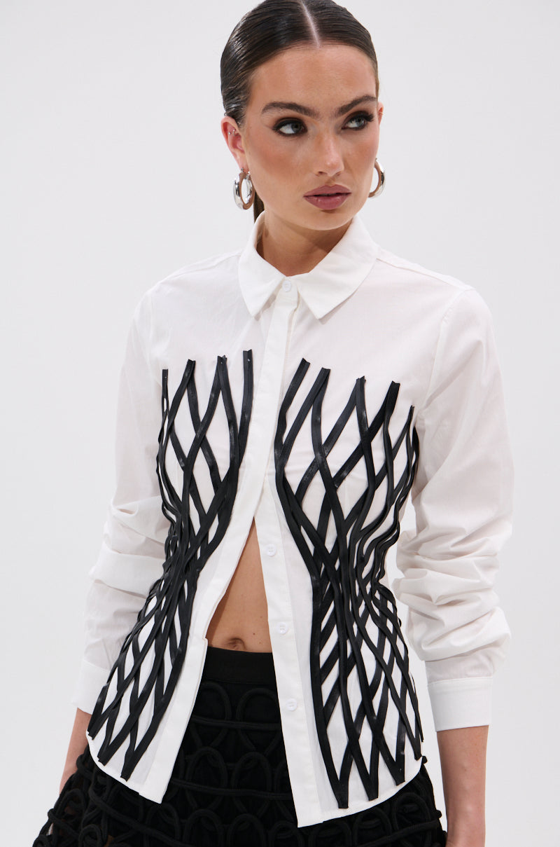 A woman with slicked-back hair models the ALL OVER THE PLACE BUTTON DOWN, a tailored shirt with bold black crisscross ribbon details on the front, paired with black patterned pants, as she looks to the side against a plain white background.