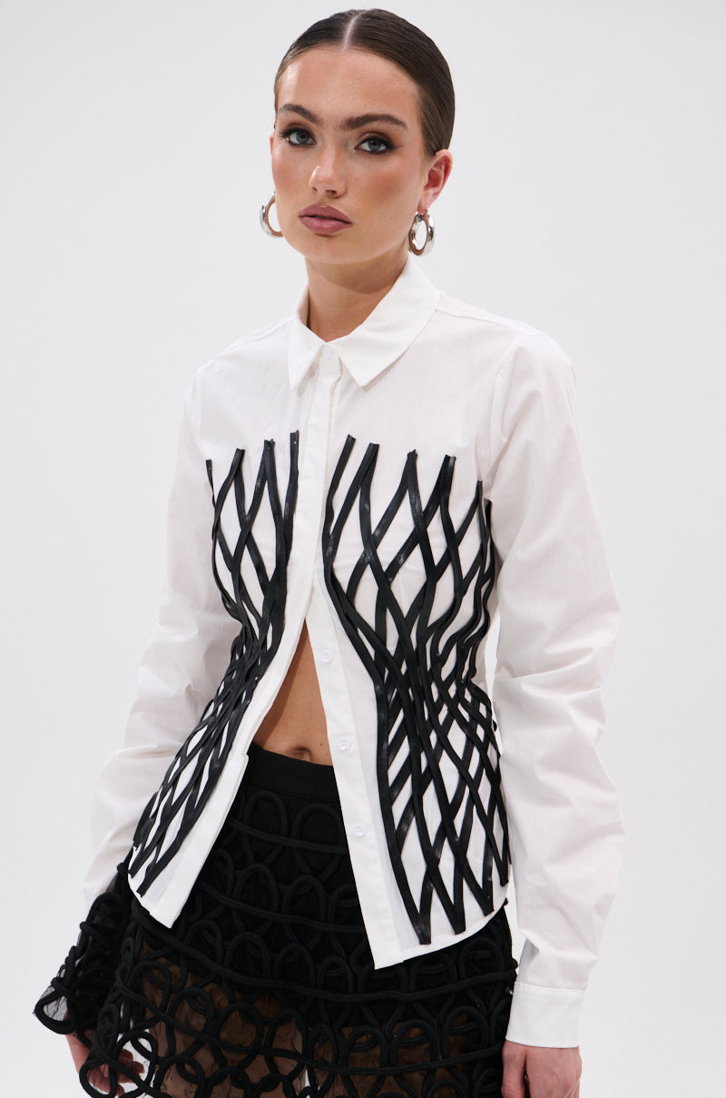 A woman models the ALL OVER THE PLACE BUTTON DOWN, a white tailored shirt with black geometric detailing, paired with a matching mesh skirt, hoop earrings, and a faux leather waistbelt against a plain white background.