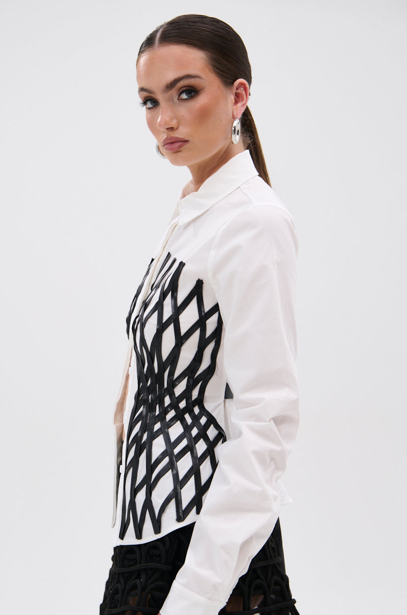 A woman with slicked-back hair wears the ALL OVER THE PLACE BUTTON DOWN, a tailored fit shirt with bold black geometric side panels. She accessorizes with silver hoop earrings and a faux leather waistbelt, looking confidently at the camera.
