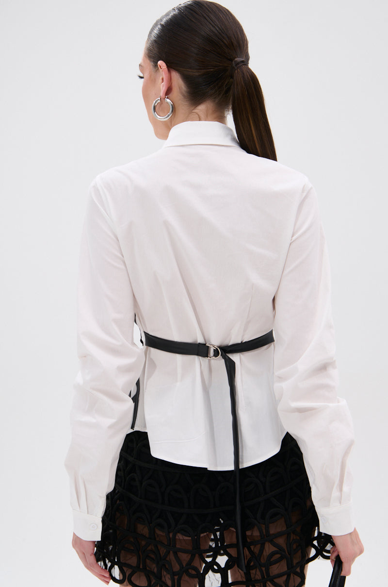 A woman with long brown hair in a ponytail wears the ALL OVER THE PLACE BUTTON DOWN, a tailored white shirt, paired with a black faux leather waistbelt, geometric-patterned mini skirt, and silver hoop earrings, seen from behind.