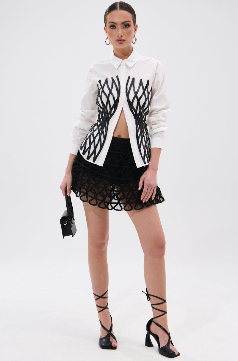 A woman wears the ALL OVER THE PLACE BUTTON DOWN, a tailored white shirt with black geometric patterns, paired with a matching mini skirt, strappy heels, hoop earrings, waistbelt, and carries a black handbag against a plain white background.