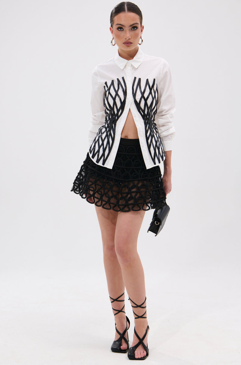 A woman stands against a white background wearing the ALL OVER THE PLACE BUTTON DOWN, a black geometric print mini skirt, strappy black heels, hoop earrings, a faux leather waistbelt, and holding a small black clutch.