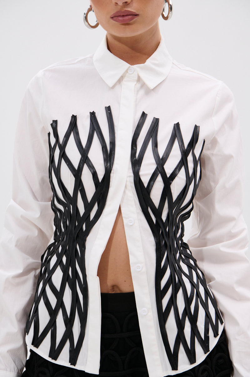 Wearing the ALL OVER THE PLACE BUTTON DOWN, featuring a tailored fit, structured collar, and black geometric lattice print across the chest and sides. The shirt is partially unbuttoned at the waist and styled with hoop earrings.