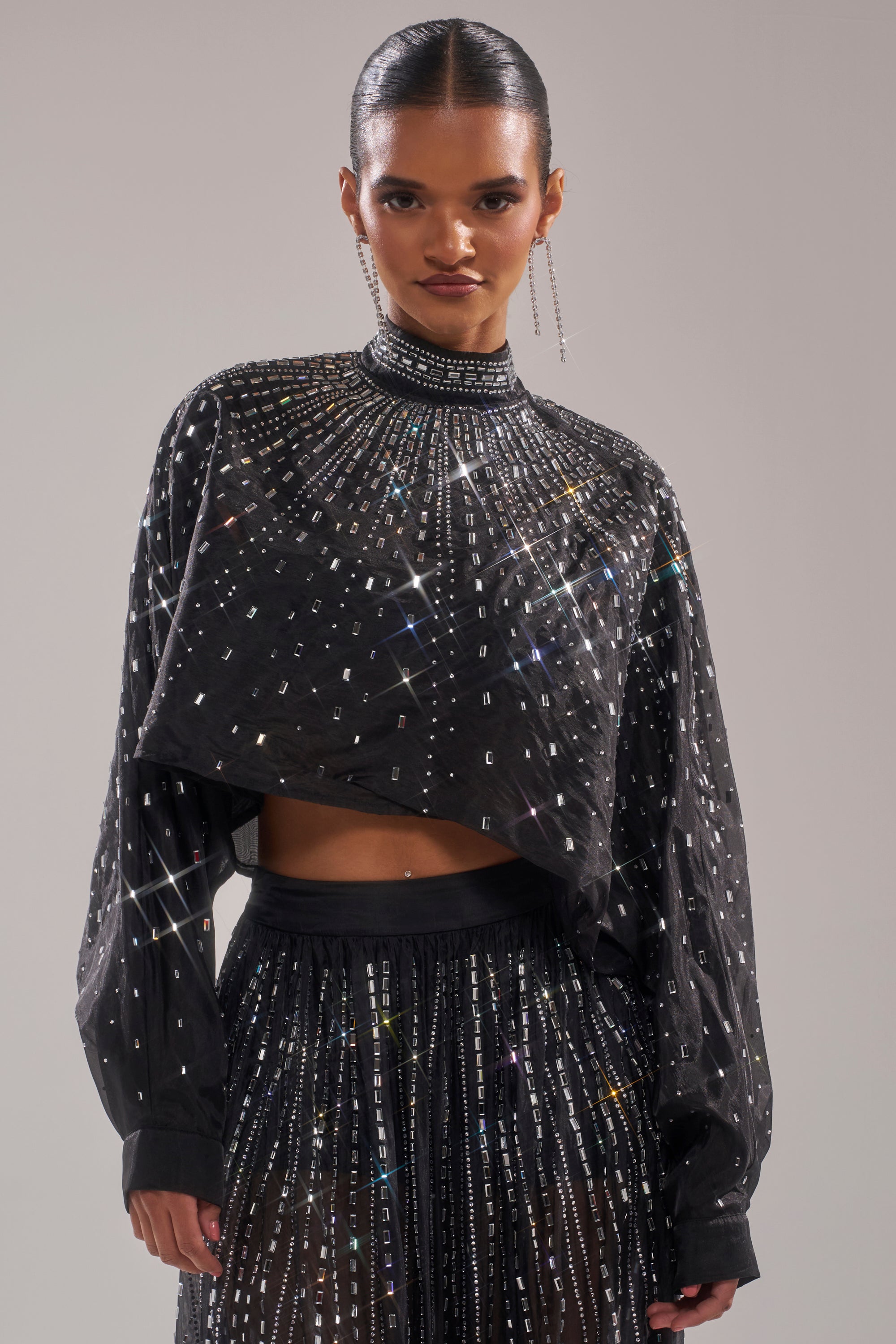 A person exudes confidence in the GLAMOROUS RHINESTONE MOCK NECK BLOUSE, styled with a sheer high-waisted skirt, long earrings, and slicked-back hair for an effortlessly chic look. 0