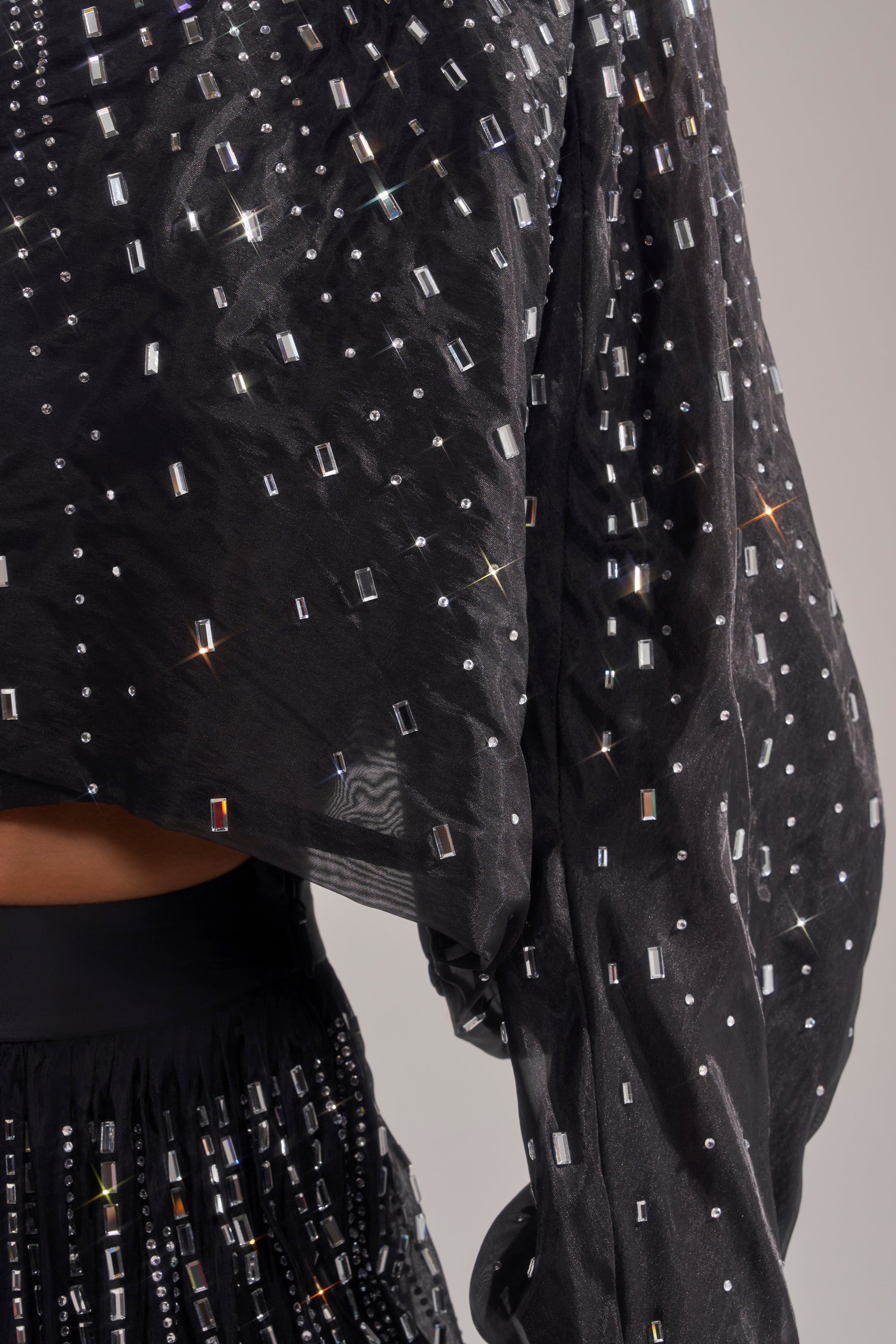 Close-up of the GLAMOROUS RHINESTONE MOCK NECK BLOUSE in sheer black, decorated with rectangular and square rhinestones; part of the sleeve and waist shown against a neutral backdrop. 9