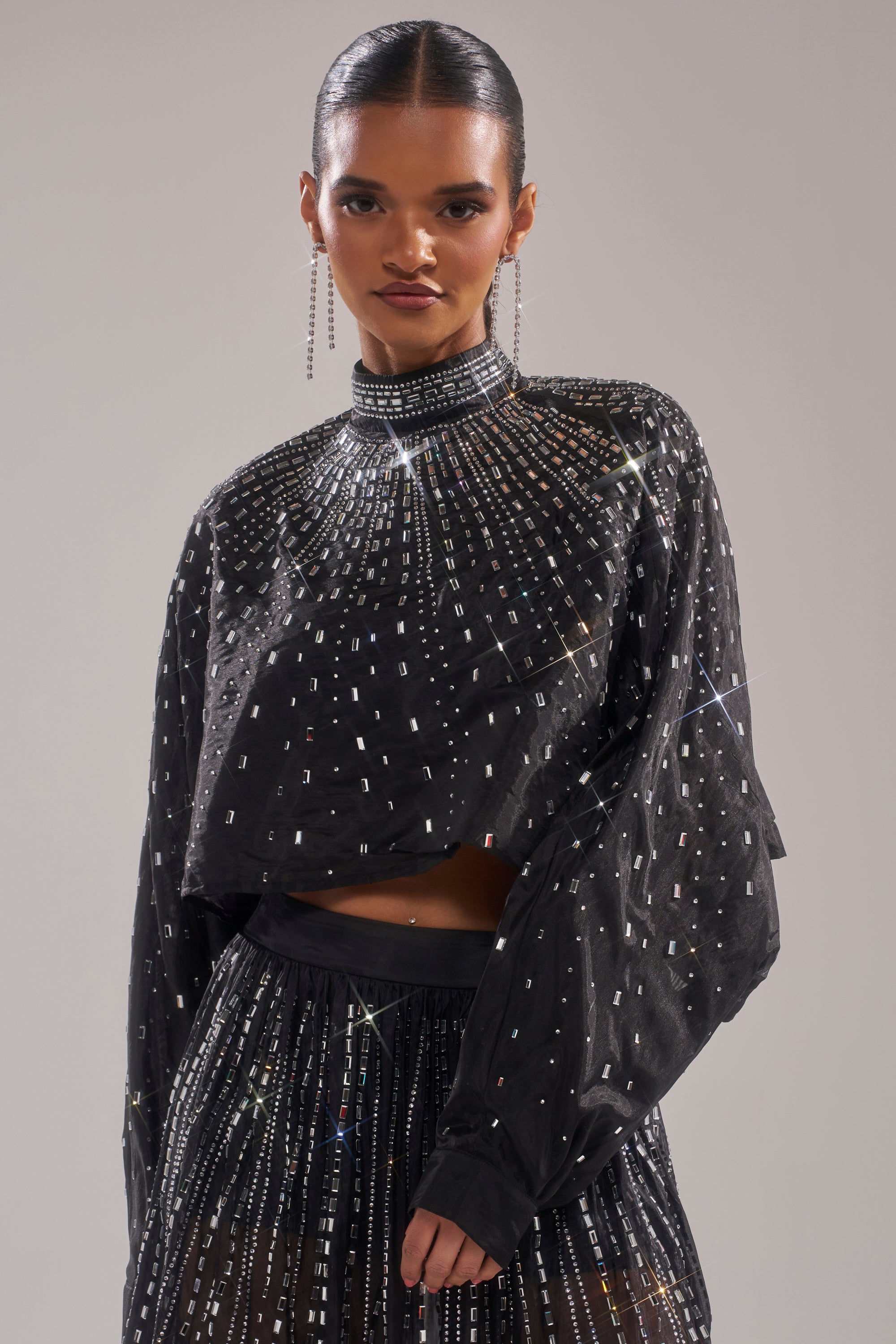 A woman with slicked-back hair wears the GLAMOROUS RHINESTONE MOCK NECK BLOUSE—a black, long-sleeve crop embellished with sparkling studs—paired with a matching skirt, long earrings, and a confident expression. 1