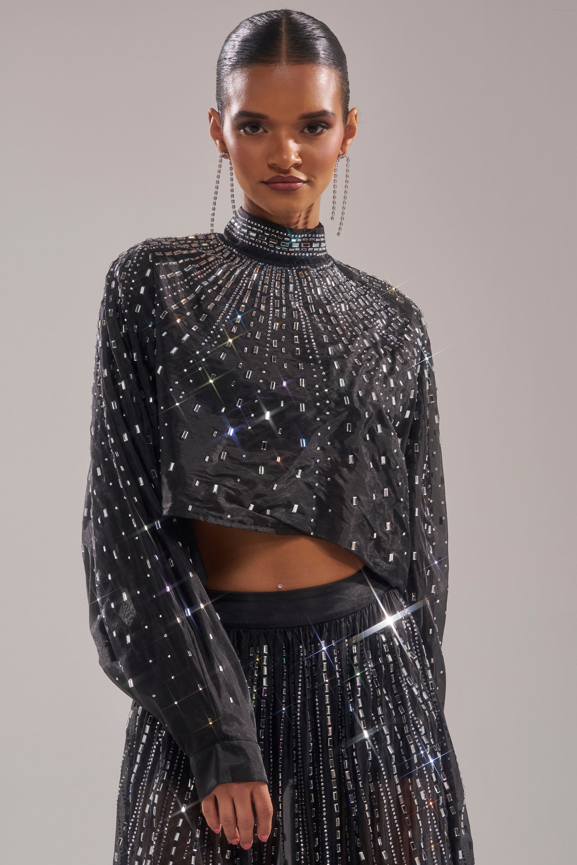 A woman poses confidently against a plain background, wearing the GLAMOROUS RHINESTONE MOCK NECK BLOUSE—black and shimmering with sequins. Her hair is slicked back and she completes the look with long, dangling earrings. 2