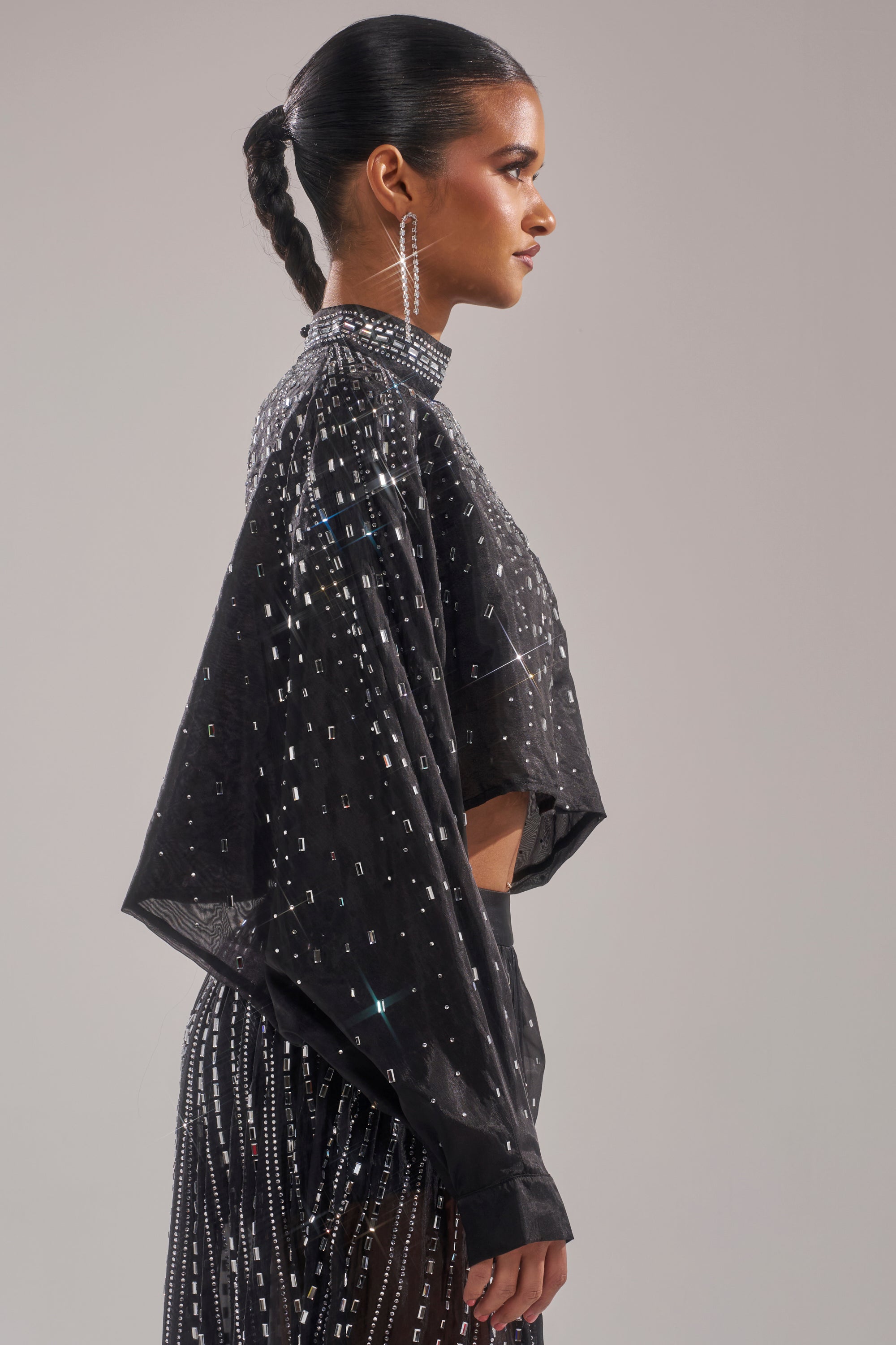 A woman in profile wears the GLAMOROUS RHINESTONE MOCK NECK BLOUSE, a sheer black top with sparkling rhinestones and long sleeves, styled with a sleek braided ponytail and long dangling earrings against a plain light background. 3