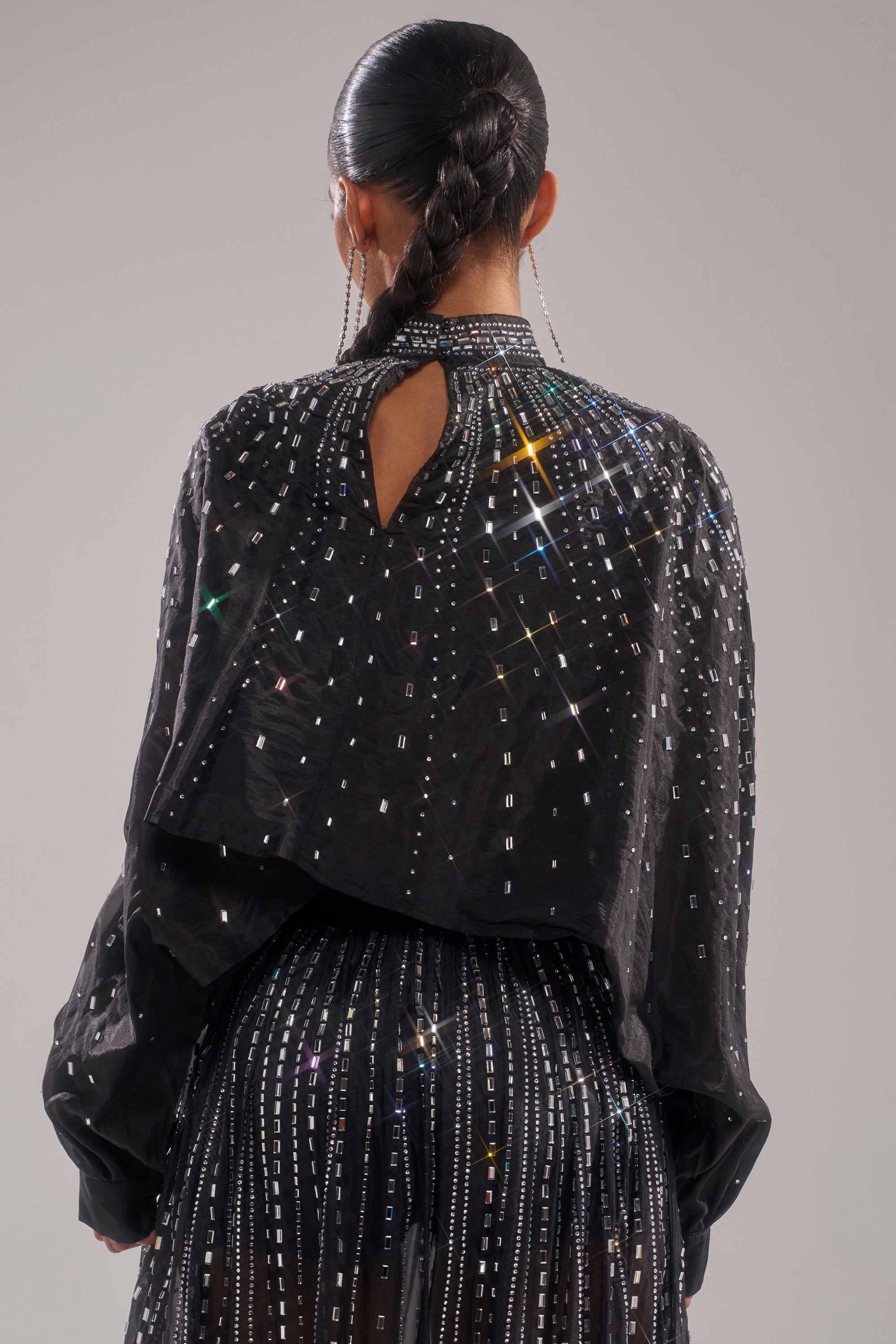A person with a long braided ponytail is seen from behind wearing the GLAMOROUS RHINESTONE MOCK NECK BLOUSE, featuring sparkling rhinestone embellishments and a back keyhole cutout against a plain light background. 4