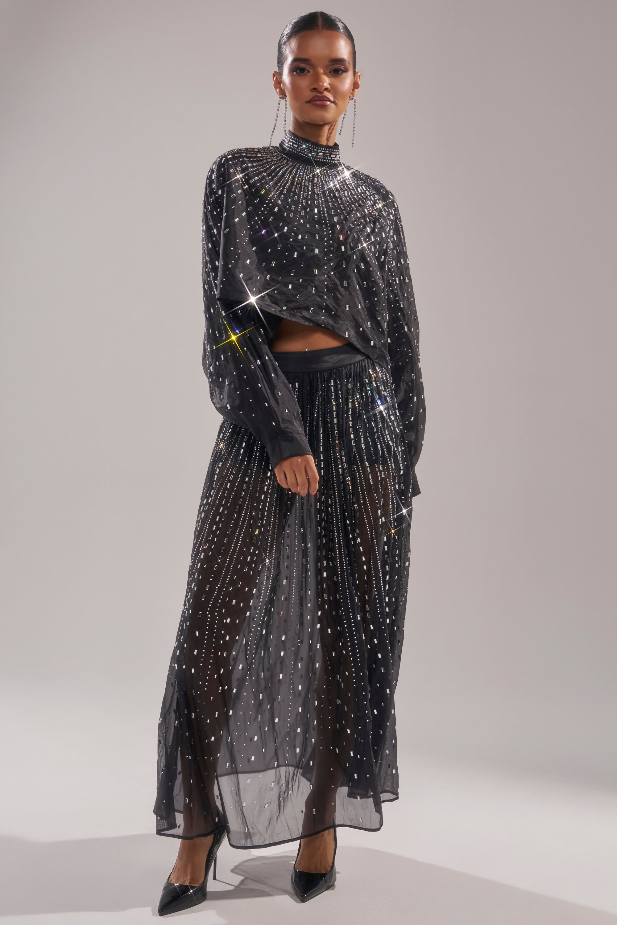 A woman models the GLAMOROUS RHINESTONE MOCK NECK BLOUSE with matching bottoms, black heels, and long earrings. Her hair is slicked back, showing off the sheer sparkling outfit and her confident look. 6